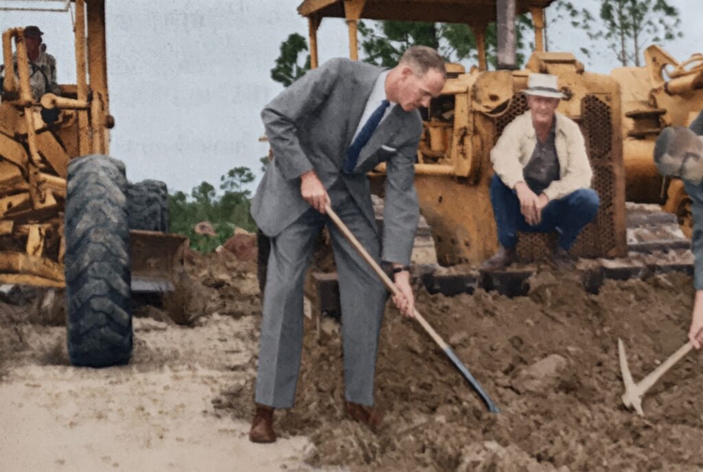 Colorized photo of a man in a gray suit turning the first shovel of dirt at the December 1962 groundbreaking for the Cape Coral Bridge, with road-building equipment behind him